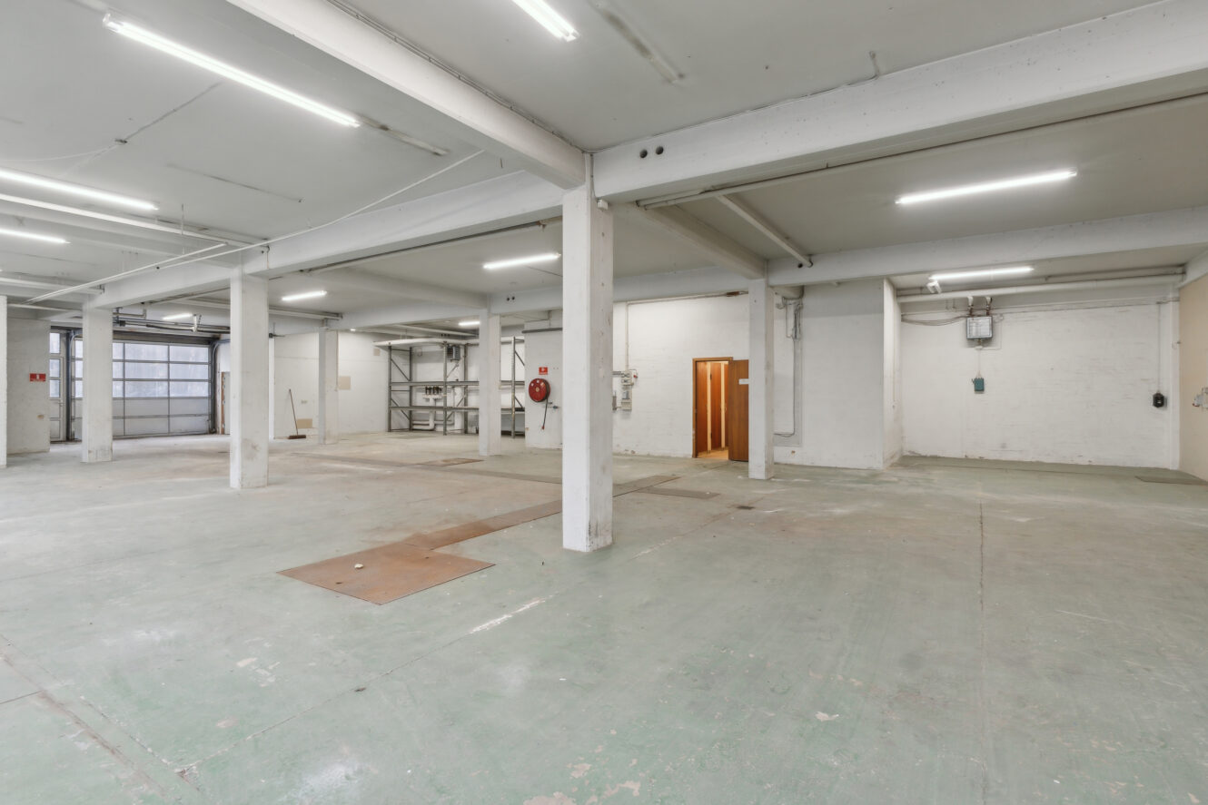 Tomt industrilager med betongulv, hvide søjler, ovenlys, hylder i baggrunden og en åben trædør på højre væg.