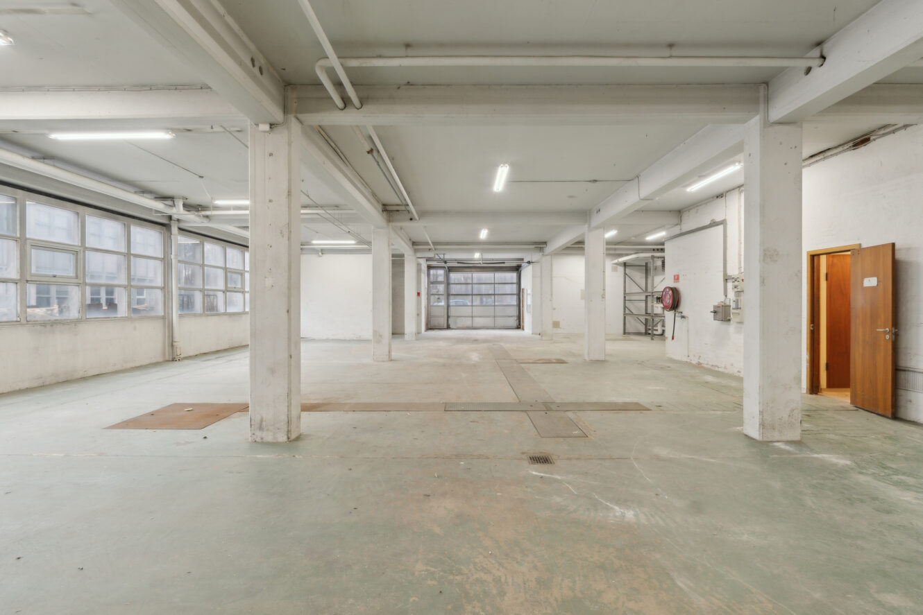 Stort, tomt industrilager med betongulv, hvide vægge, store vinduer i den ene side, lysstofrør og en åben trædør.