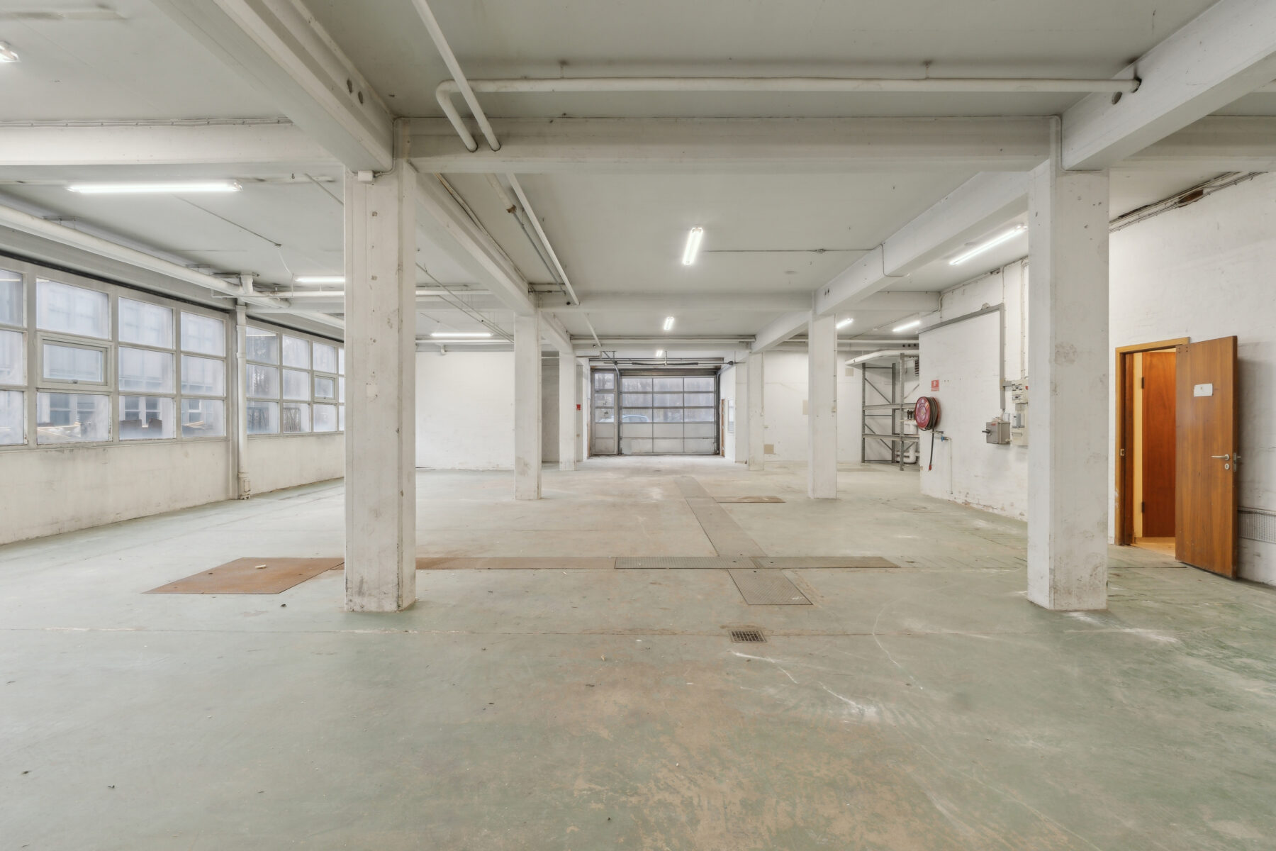 Stort, tomt industrilager med betongulv, hvide vægge, store vinduer i den ene side, lysstofrør og en åben trædør.