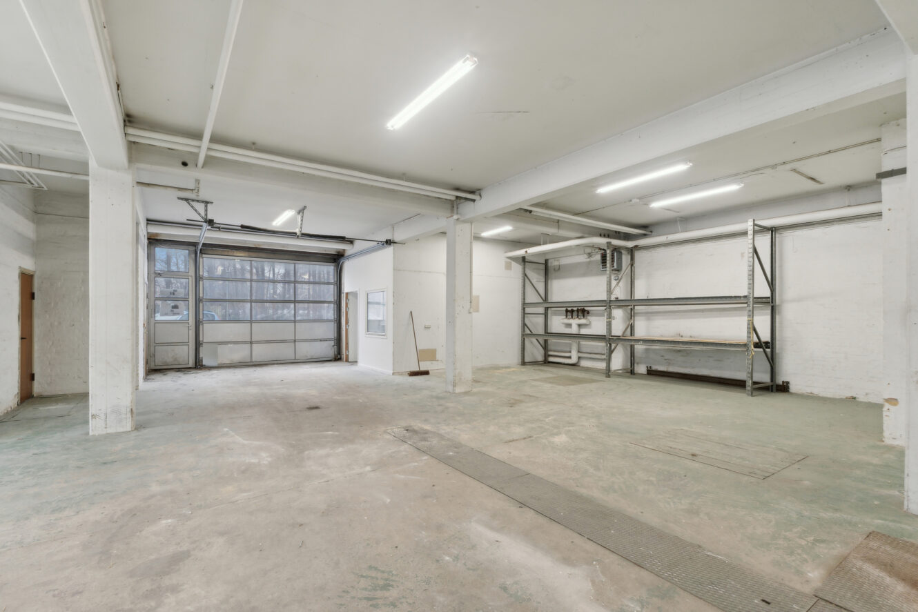 Stort, tomt industrilager med betongulv, hvide vægge, ovenlys, metalreoler og en stor garageport.