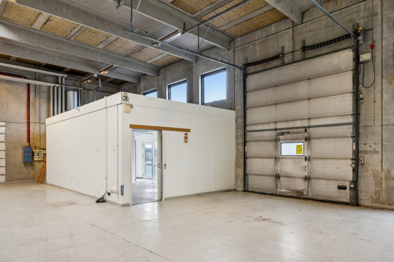 Interiør i et tomt industrilager med et lille hvidt kontorlokale, højt til loftet, stor garageport og flere vinduer, der giver naturligt lys.