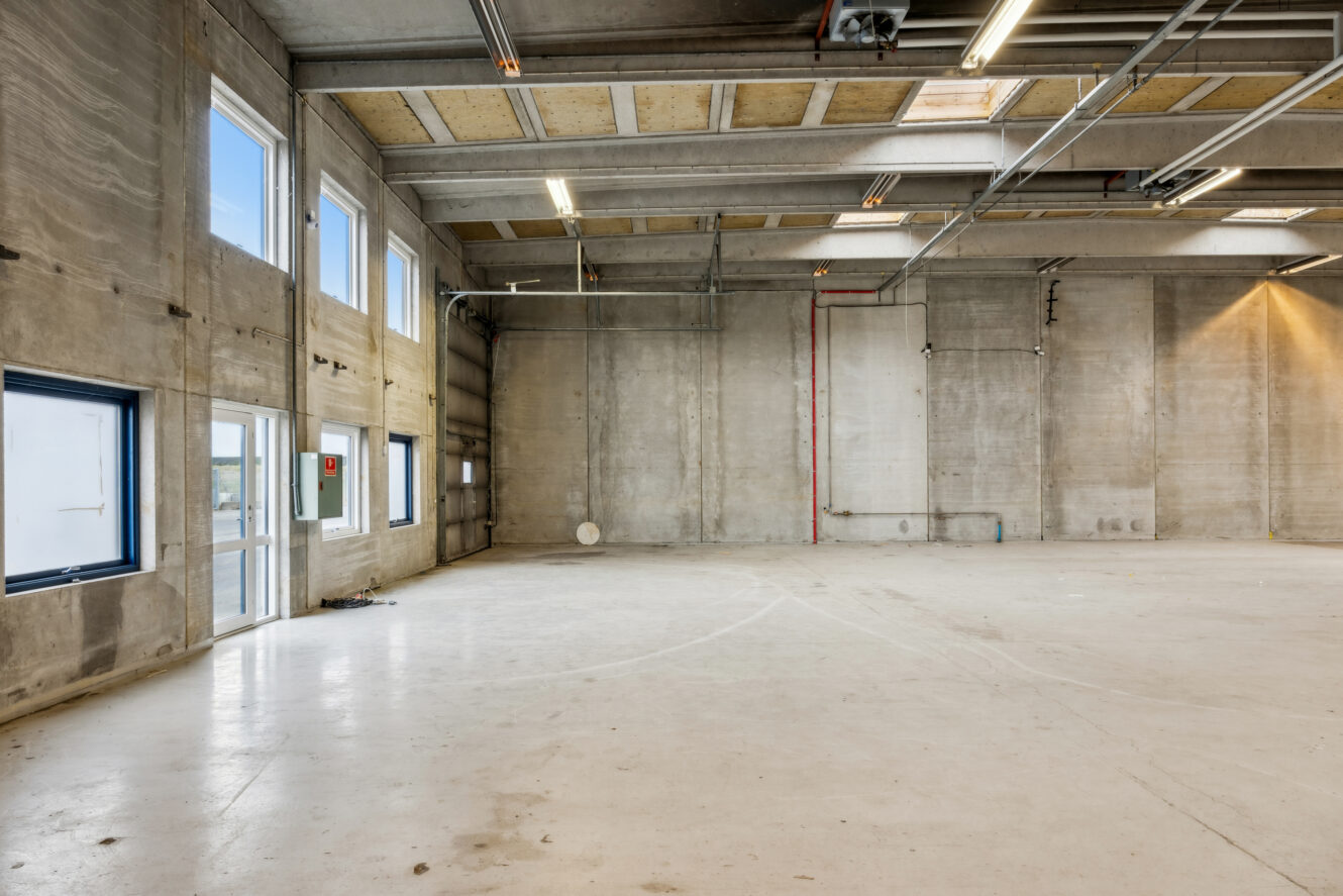 Stort tomt industrilager med betonvægge, højt til loftet, store vinduer og synlige rør og ledninger.