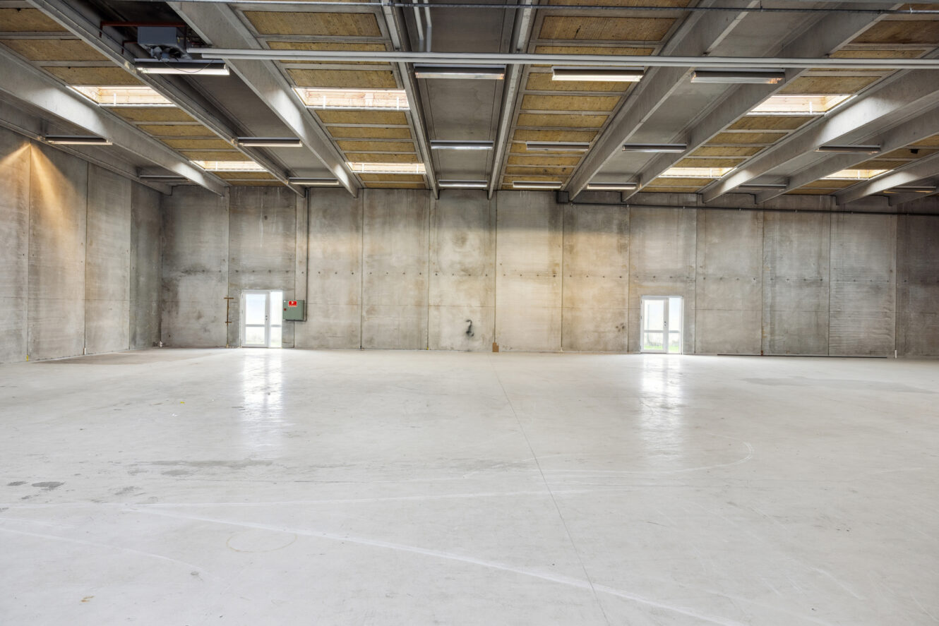 Et stort, tomt industrilokale med betonvægge, højt til loftet, ovenlys og to små døre langs bagvæggen.