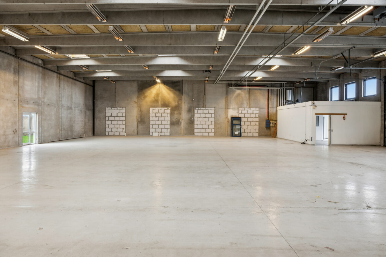 Stor tom lagerhal med betonvægge, højt til loftet, synlige bjælker, lysstofrør og en lille hvid kontorsektion i hjørnet.