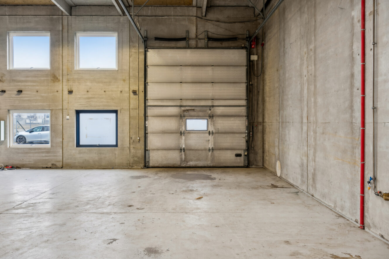 Tom industrigarage med betonvægge, stor rulleport, tre vinduer og synlige loftsbjælker. Et rødt rør løber lodret langs den højre væg.