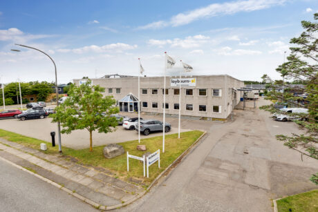 En toetagers erhvervsbygning med Laybourn-skiltning og -flag, omgivet af en parkeringsplads med parkerede biler og træer, under en delvis overskyet himmel.