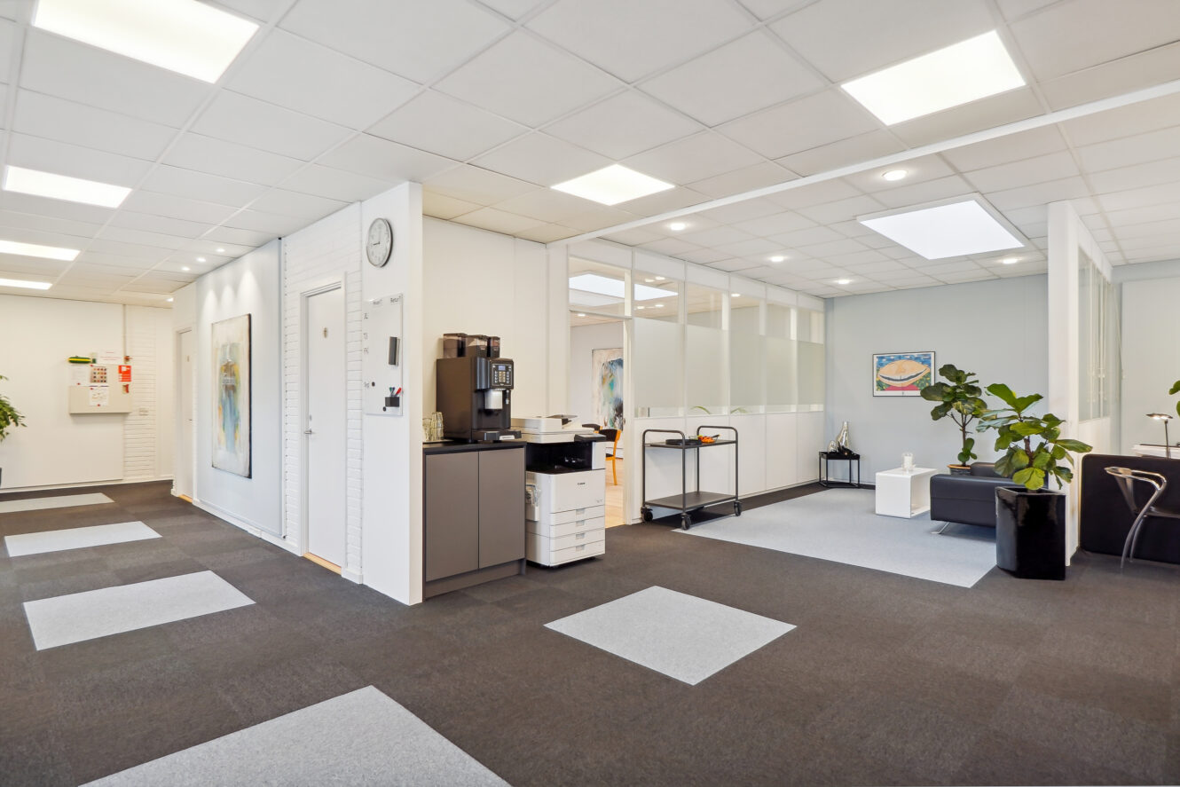 Moderne kontor med hvide vægge, glasskillevægge, kaffemaskine, printer, potteplanter og minimalistiske møbler under stærke loftslamper.