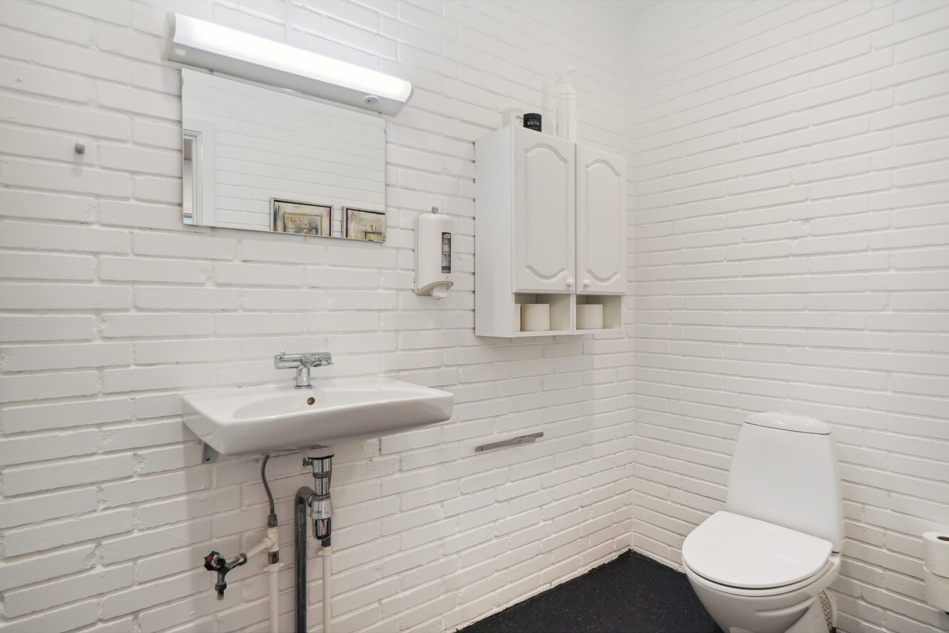 Et lille badeværelse med hvide murstensvægge, en væghængt vask, et spejl, et toilet og et hvidt skab over toilettet.