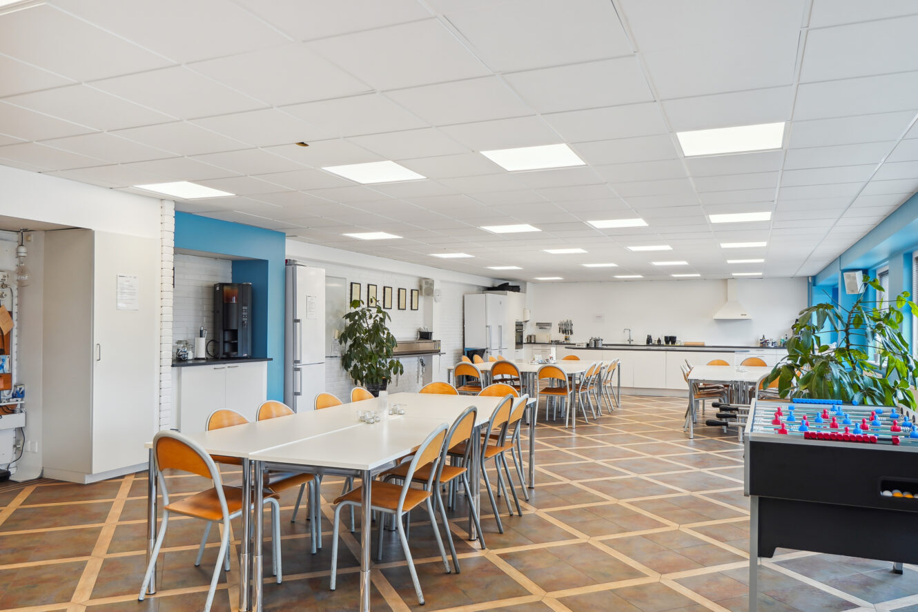 Et rummeligt, moderne pauserum med borde og stole, et køkkenområde, potteplanter og et bordfodboldbord, alt sammen oplyst af loftslamper.