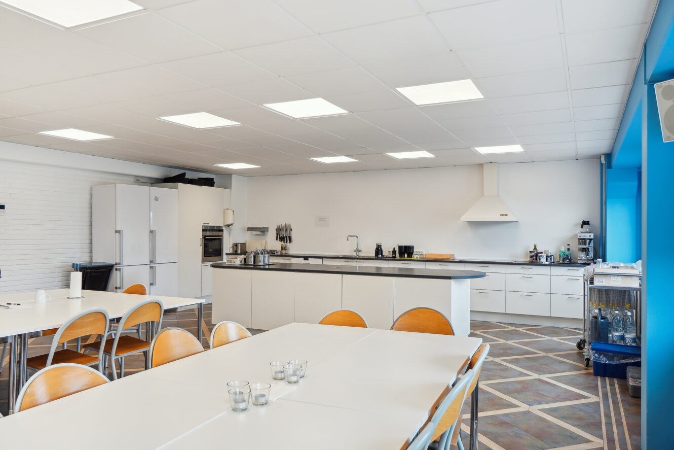 Et rummeligt, moderne køkken med hvide skabe, træstole, borde, stort køleskab, ovn og køkkenmaskiner under lyse loftslamper.