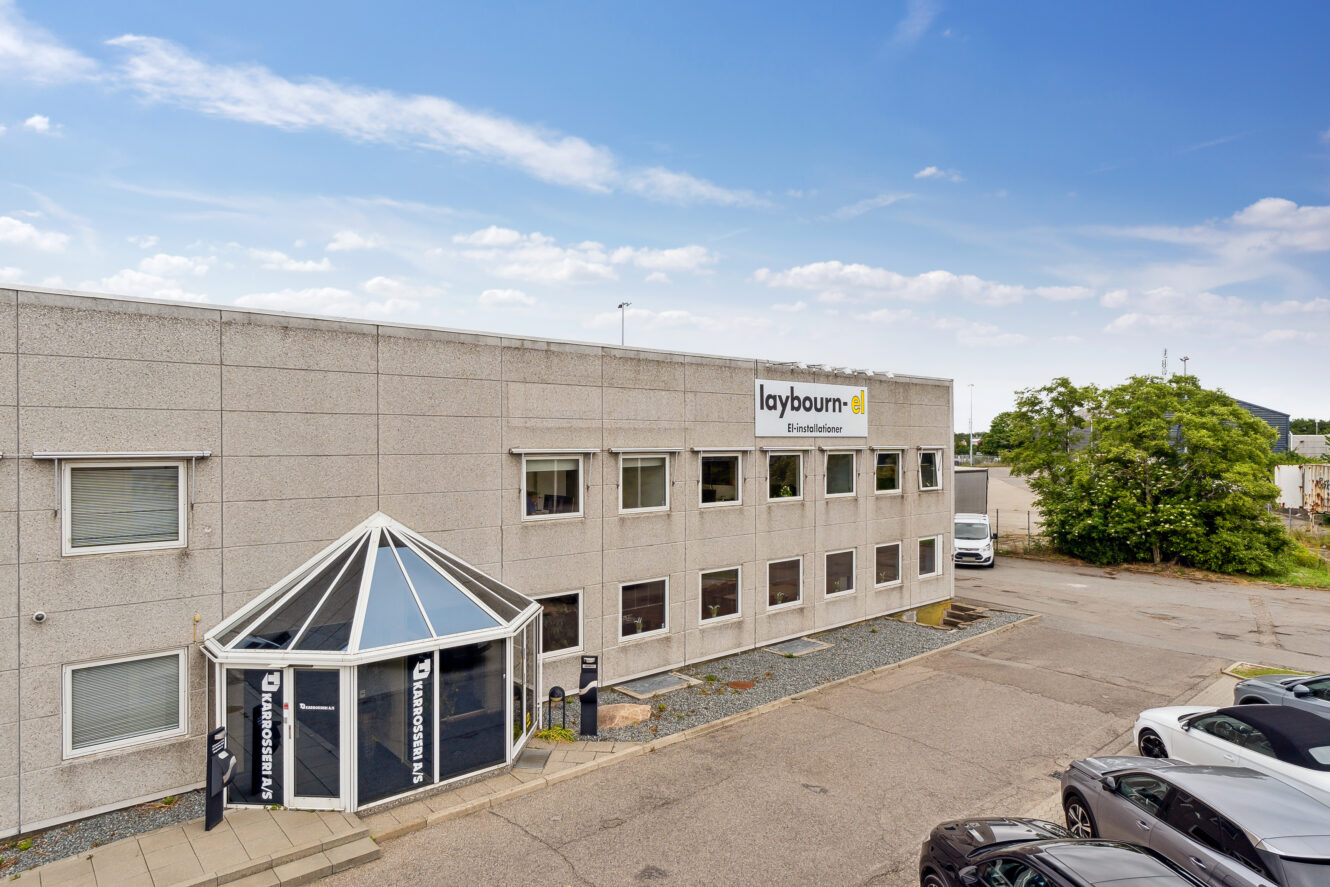 En toetagers grå erhvervsbygning med en glasindgang og et skilt med teksten Laybourn Frøavlshuset på facaden; parkerede biler og træer ses i nærheden under en blå himmel.