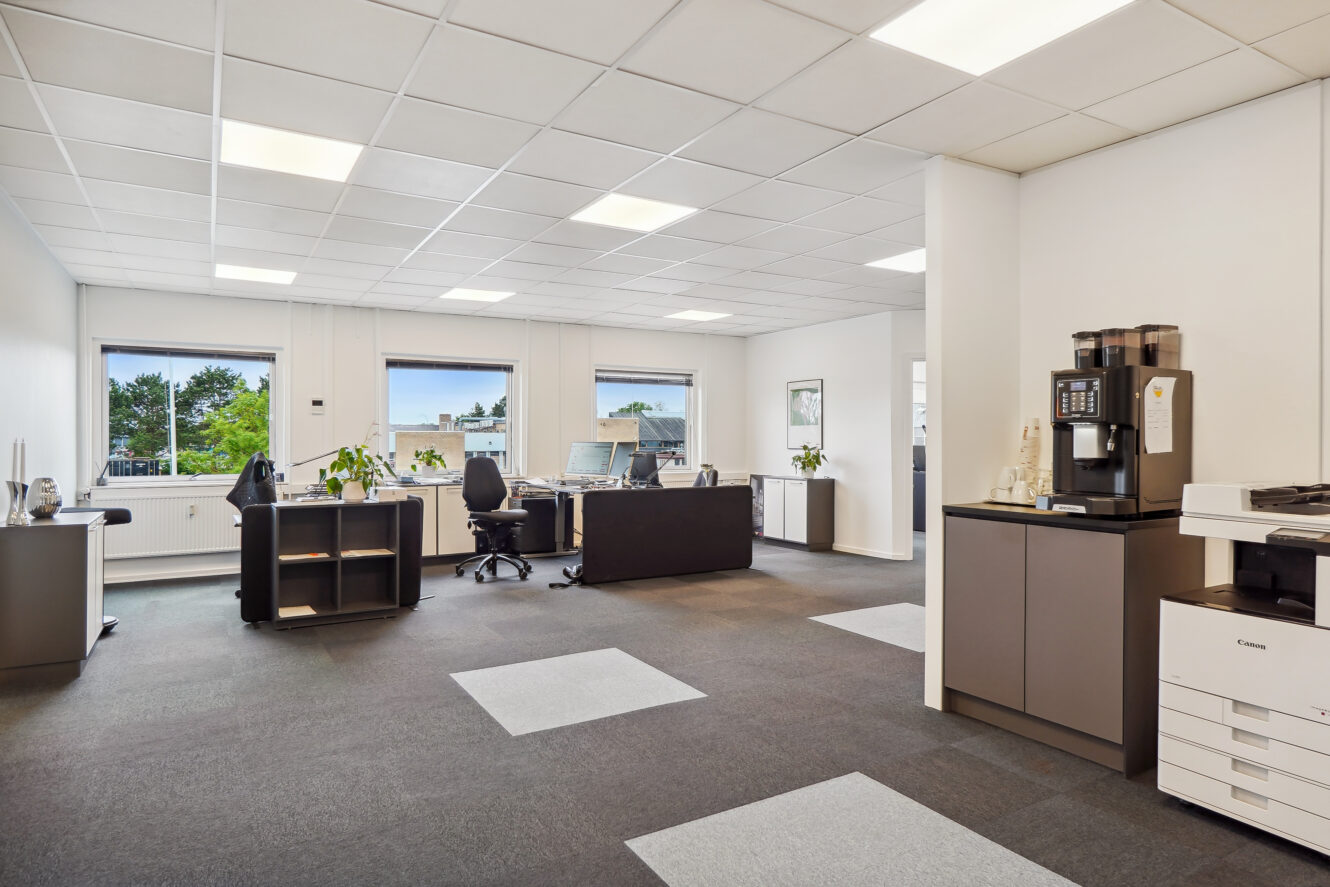 Moderne kontor med skriveborde, stole, computere, potteplanter, kaffemaskine og printer. Store vinduer giver naturligt lys og udsigt til det fri.