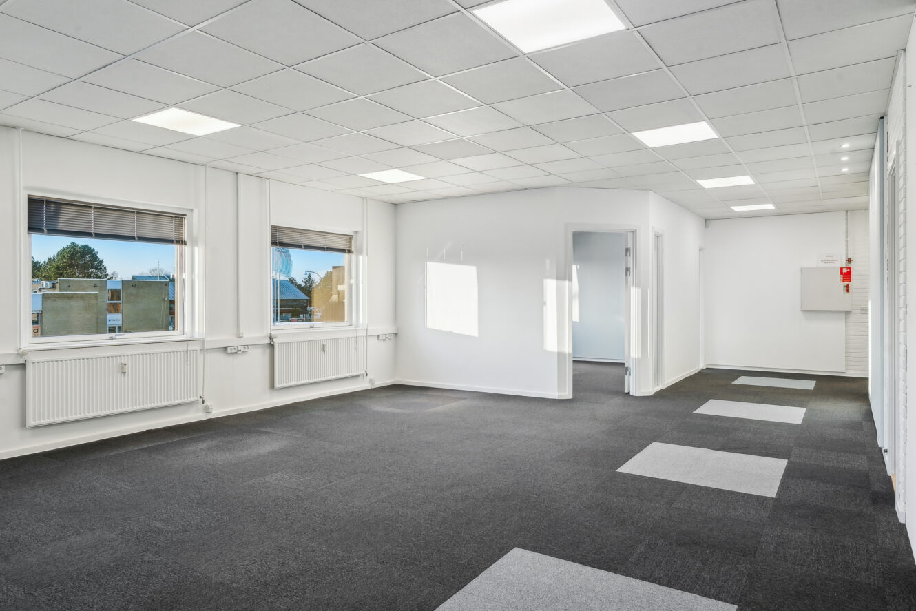 Tomt kontorlokale med grå tæppefliser, hvide vægge, store vinduer til venstre, lysstofrør i loftet og et lille rum eller kontor i den fjerneste ende.