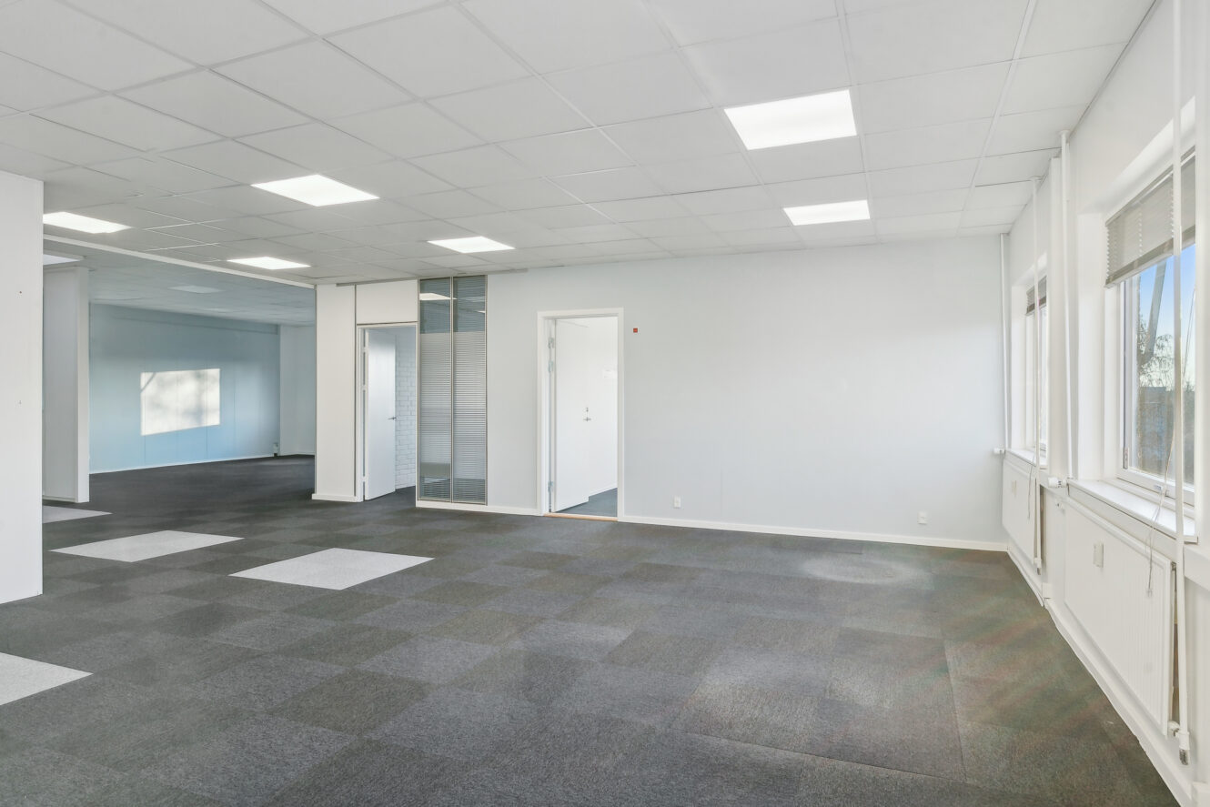 Tomt kontorlokale med hvide vægge, nedhængt loft, store vinduer og gulvtæppe i ternet mønster.