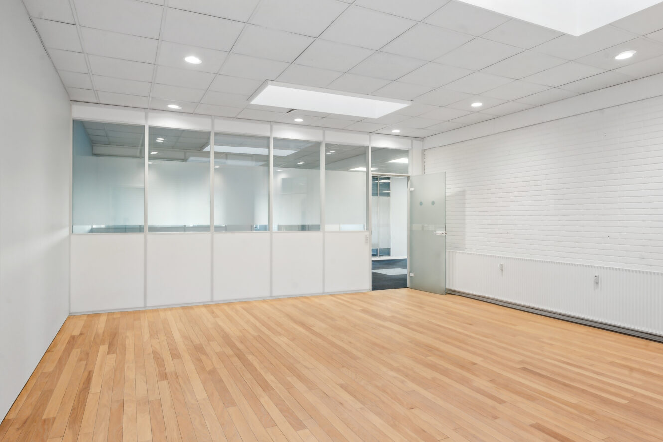 Tomt kontorlokale med glasskillevæg, hvide loftsplader, indbygget belysning, trægulv og en dør til højre.