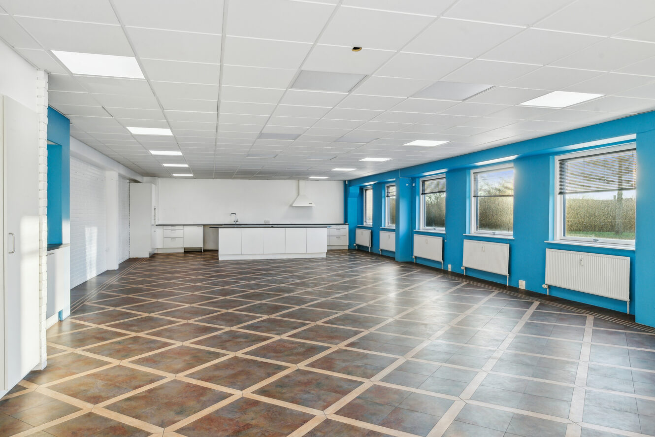 Rummeligt, tomt rum med klinkegulv, blå og hvide vægge, store vinduer og et køkkenområde i den anden ende.