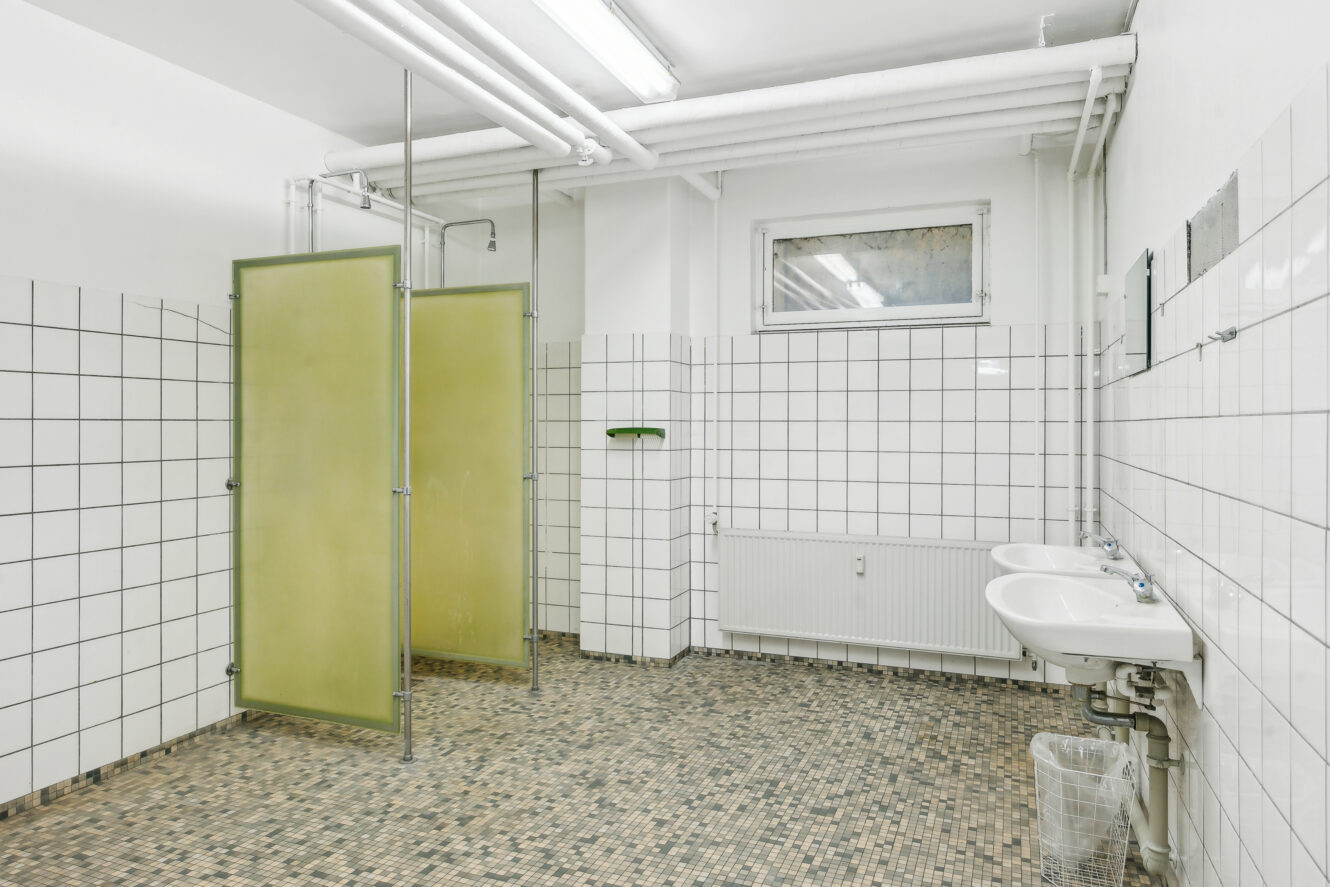 Et offentligt bruserum med flisebelagte vægge og gulv, to håndvaske, en papirkurv og to brusekabiner med skillevægge af matteret glas.