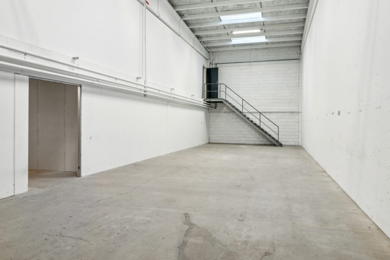 Tomt industrilager med betongulv, hvide vægge, højt til loftet, ovenlysvinduer, en åben døråbning og en metaltrappe, der fører op til en platform bagved.