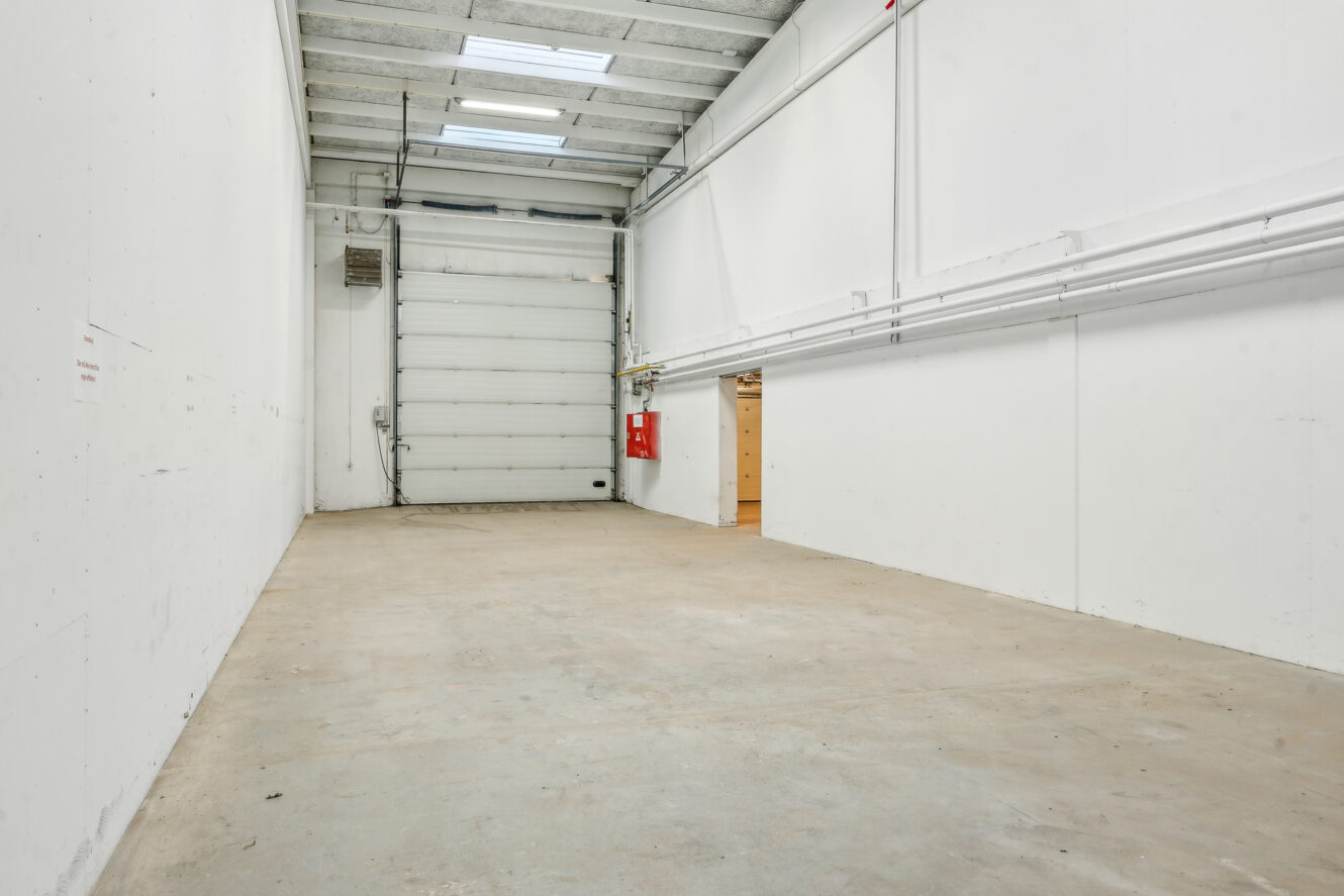 Tomt industrilager med betongulv, hvide vægge, højt til loftet, ovenlysvinduer, en stor rulleport, brandslukker og synlige rør langs højre væg.