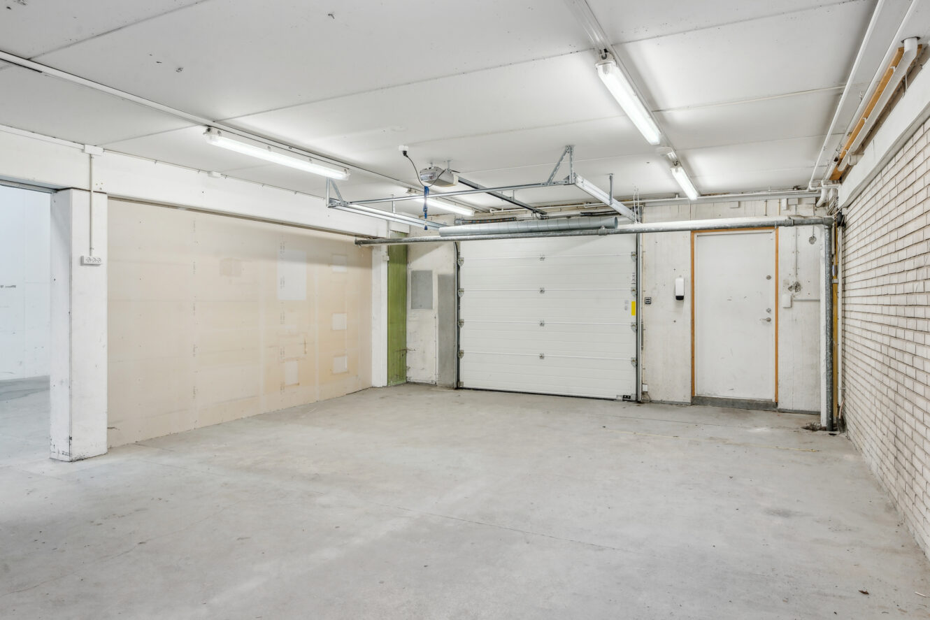 Tom garage indvendigt med betongulv, hvide vægge, lysstofrør, en lukket garageport og en sideindgangsdør.