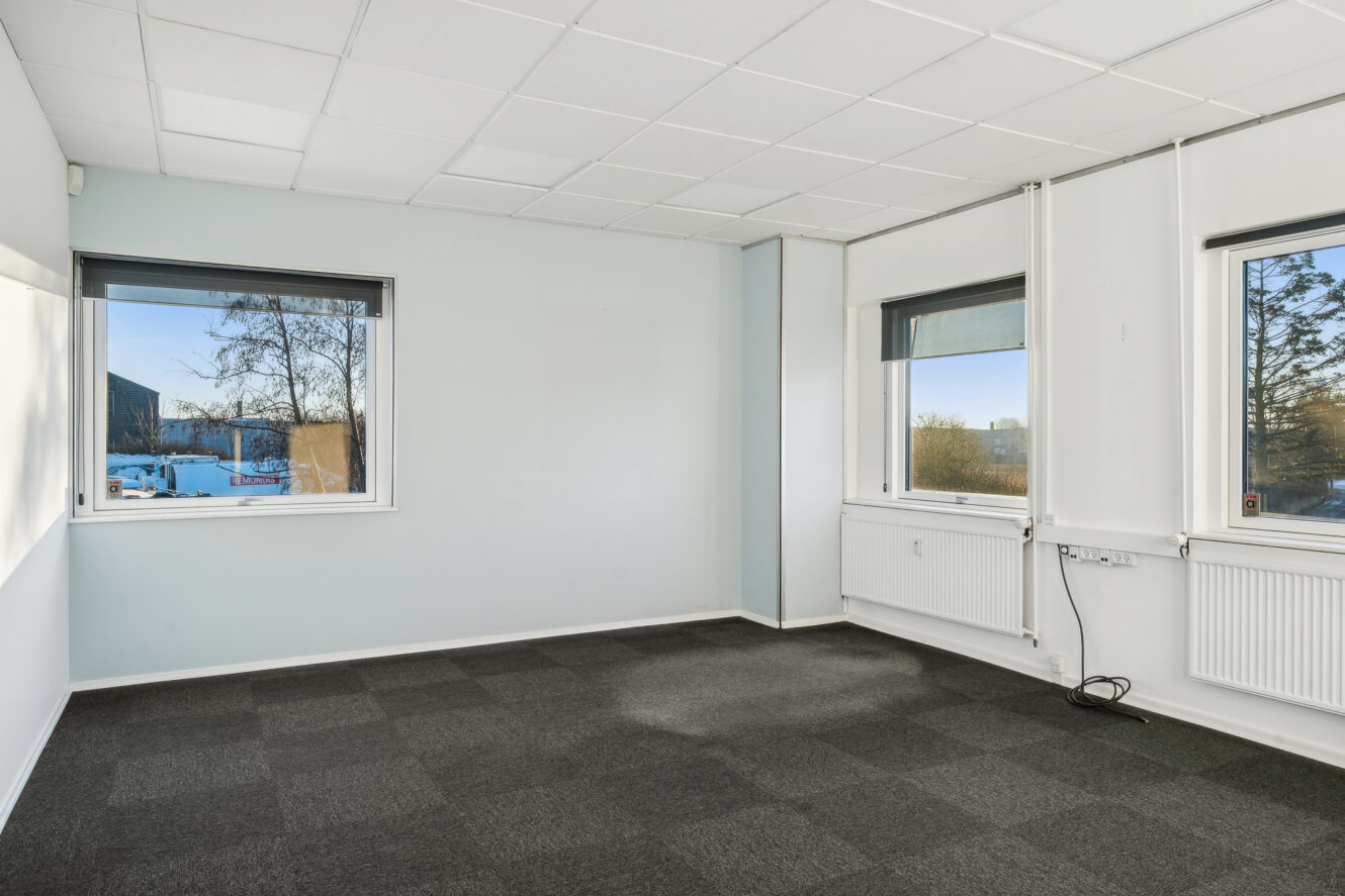 Tomt kontorlokale med hvide vægge, mørkt gulvtæppe og tre vinduer, der lukker naturligt lys ind. Radiatorer og stikkontakter er synlige langs væggene.
