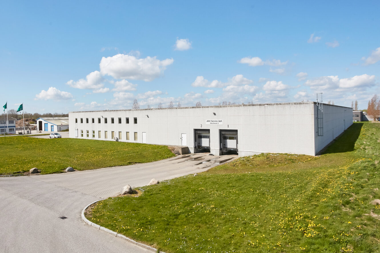 Et stort, hvidt industrilager med to læsseramper og flere vinduer, omgivet af græs og en asfalteret indkørsel under en blå himmel med spredte skyer.