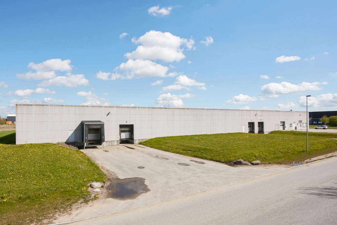 Et stort, vinduesløst industrilager med læsseramper og en asfalteret indkørsel, omgivet af græs under en blå himmel med skyer.