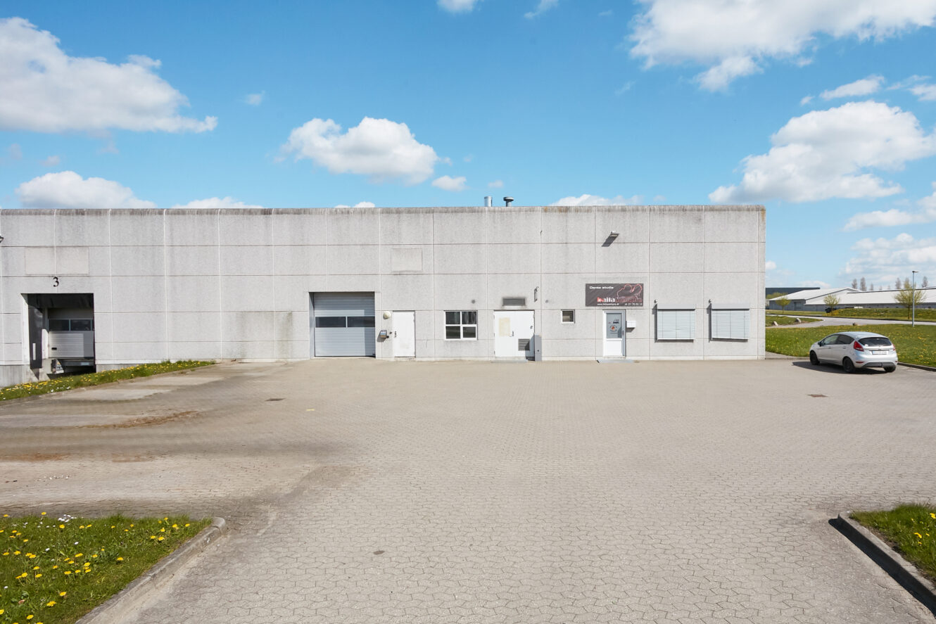 En almindelig industribygning med et gråt ydre, flere døre og vinduer, en parkeret hvid bil og en klar himmel med spredte skyer.