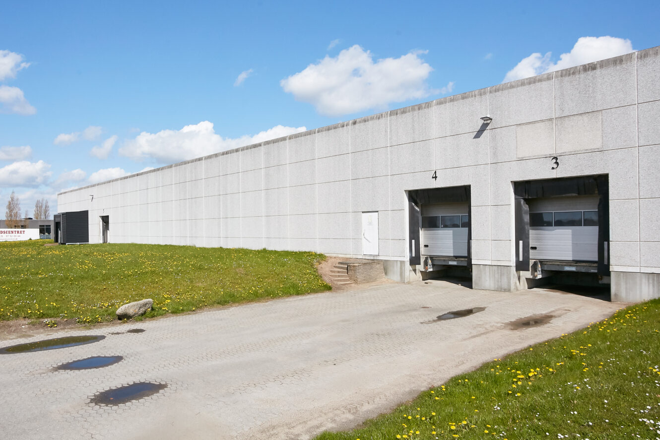 En lagerbygning i beton med læsseramper nummer 3 og 4, lastbiltrailere, der er bakket ind, og et græsareal med vandpytter i forgrunden under en delvist overskyet himmel.