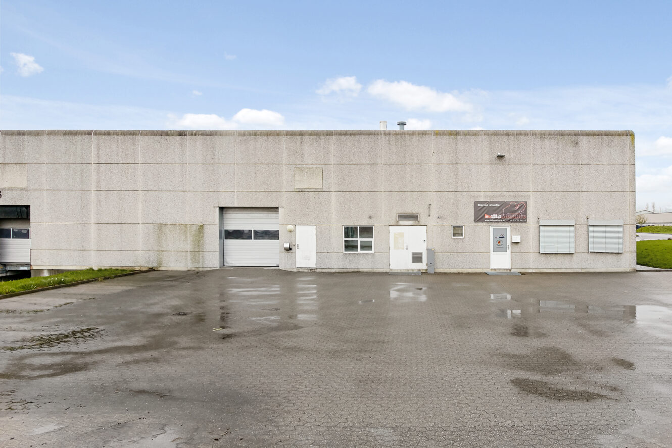 En stor, almindelig industribygning med en lukket garageport, hvide indgangsdøre, små vinduer og en tom, våd asfaltplads foran.