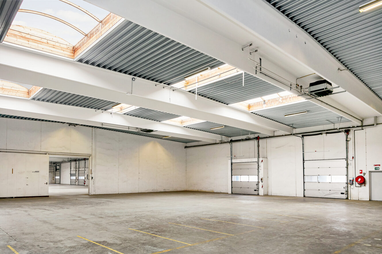 Stor tom lagerhal med betongulv, højt til loftet, ovenlys og flere store garageporte. Markerede parkeringspladser er synlige på gulvet.