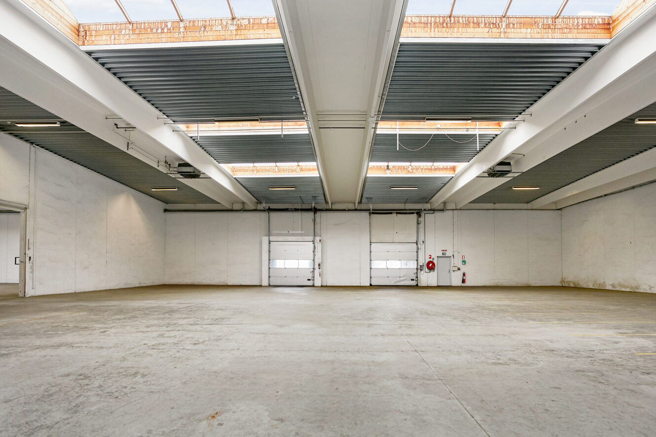 Rummelig tom lagerhal med betongulv, hvide vægge, højt til loftet, ovenlys og store lukkede garageporte på bagvæggen.