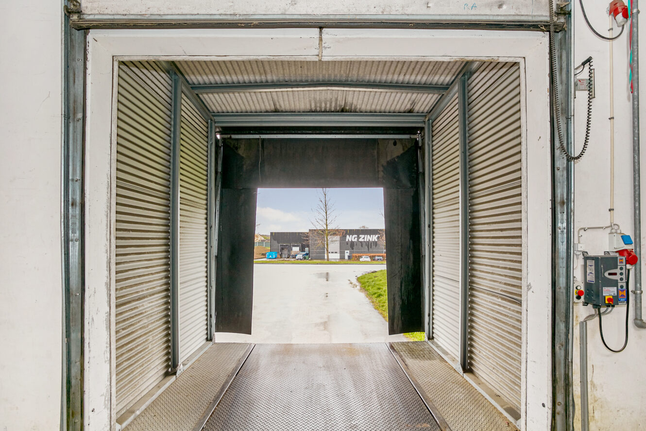 Udsigt inde fra en åben læsserampe med rulleporte i metal med udsigt til et industriområde og en bygning mærket NG ZINK.