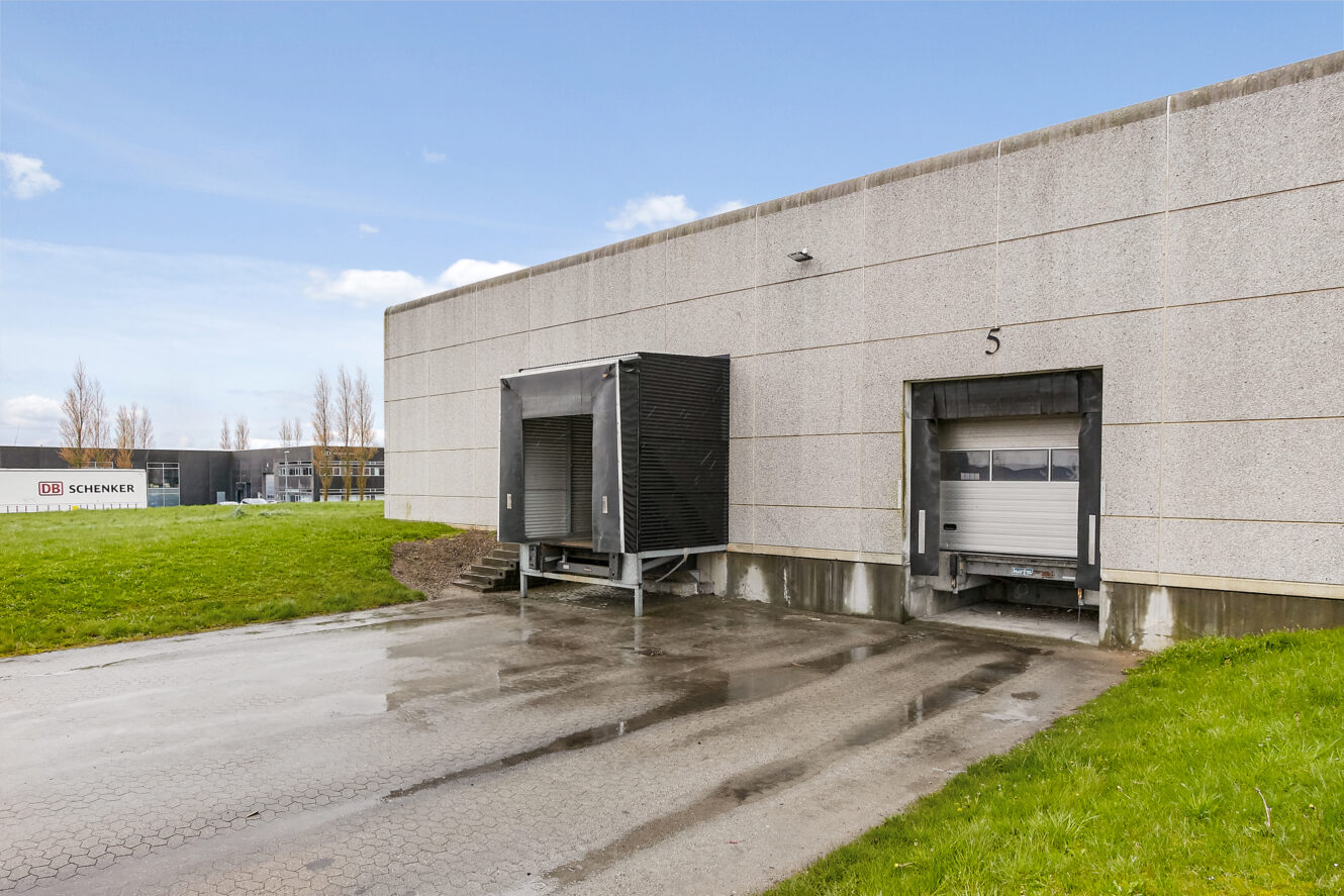 To læsseramper med lukkede ledhejseporte på et industrilager i beton, nummer 5, med en DB Schenker-bygning i baggrunden.