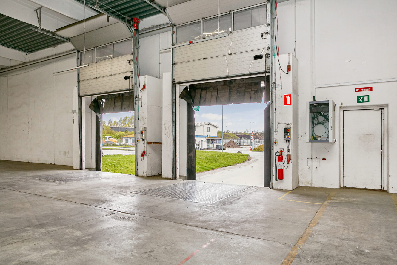 Interiør af en lagerbygning med to åbne læsseramper, brandsikkerhedsudstyr, eltavle og et udgangsskilt på væggen.