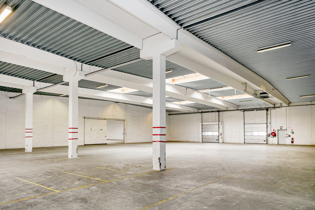 Tomt industrielt lagerinteriør med betongulv, hvide vægge, støttesøjler, ovenlys og store læssedøre.