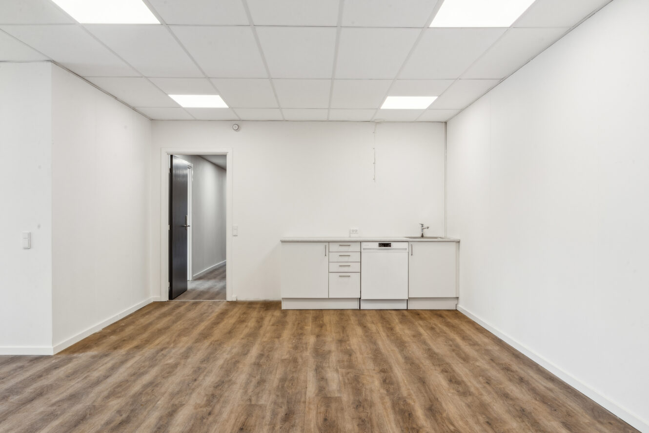 Tomt rum med trægulv, hvide vægge og et lille tekøkken med skabe, vask og opvaskemaskine på den fjerneste væg; en åben dør fører ud til en gang.