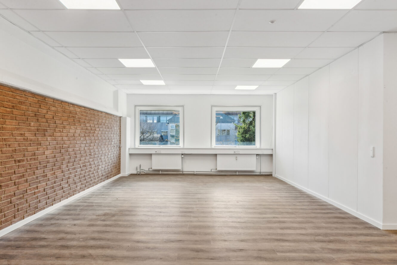 Tomt rum med træmønstret gulv, en murstensvæg til venstre, hvide vægge, to vinduer og et hvidt nedhængt loft med indbygget belysning.