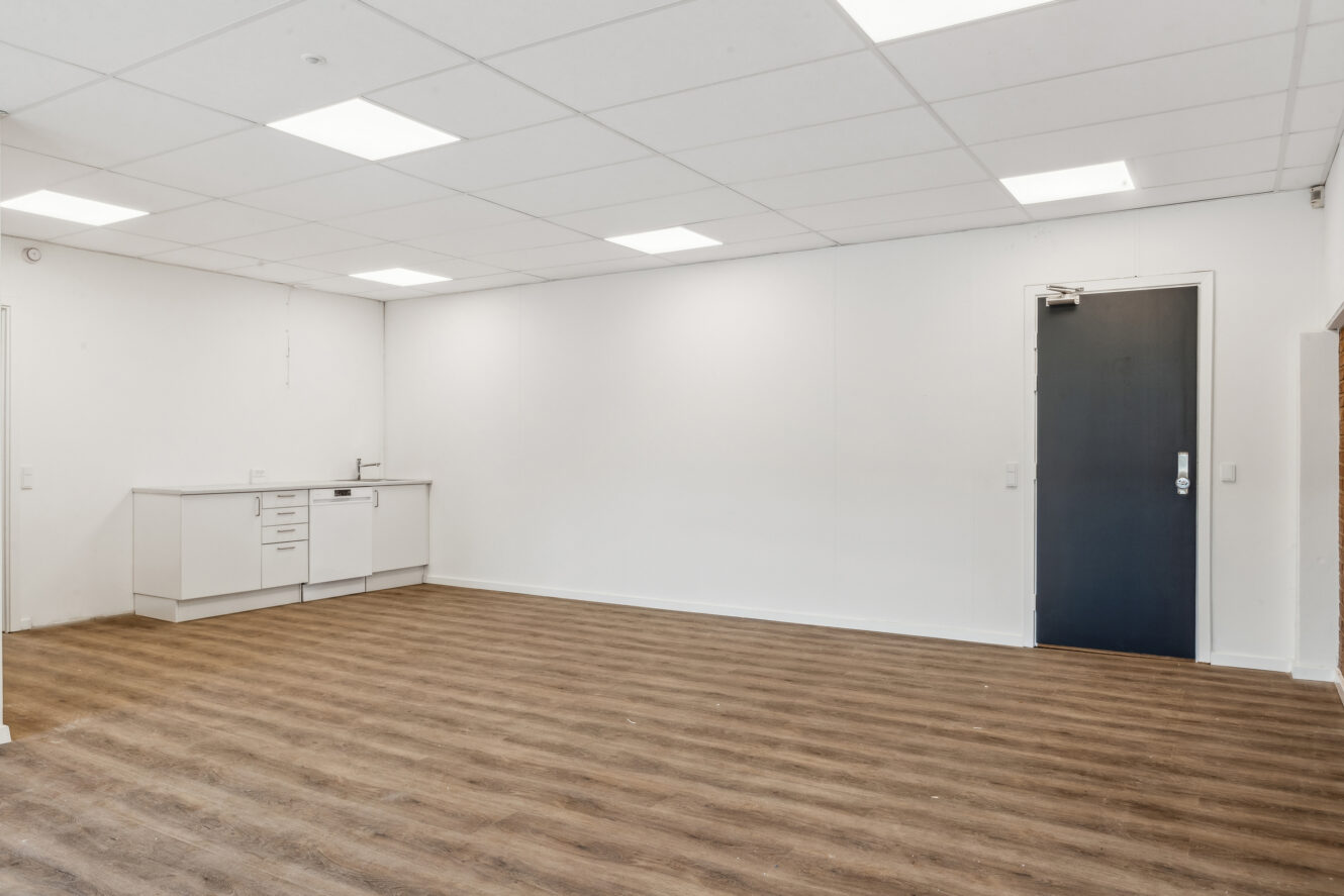 Tomt rum med trægulv, hvide vægge, en mørk dør og et lille tekøkken i hjørnet under et loft med indbygget belysning.