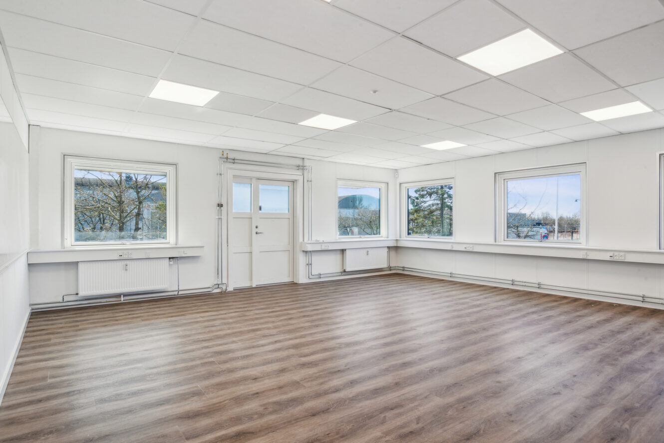Lyst, tomt rum med hvide vægge, store vinduer, en dør og trælaminatgulv. Loftet har indbyggede lysstofrør.