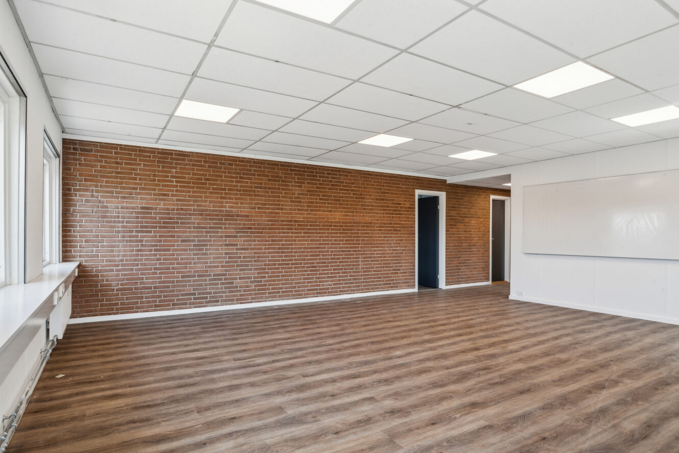 Tomt rum med træmønstret gulv, hvide loftsplader, en murstensvæg, to døråbninger, store vinduer og en whiteboardtavle på højre væg.