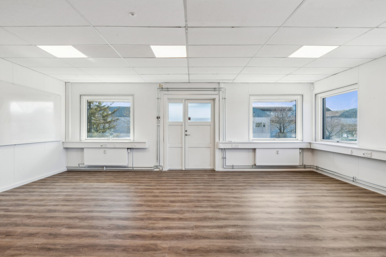 Et tomt rum med trægulv, hvide vægge, et whiteboard til venstre, tre vinduer og en dør i midten, der lukker naturligt lys ind.