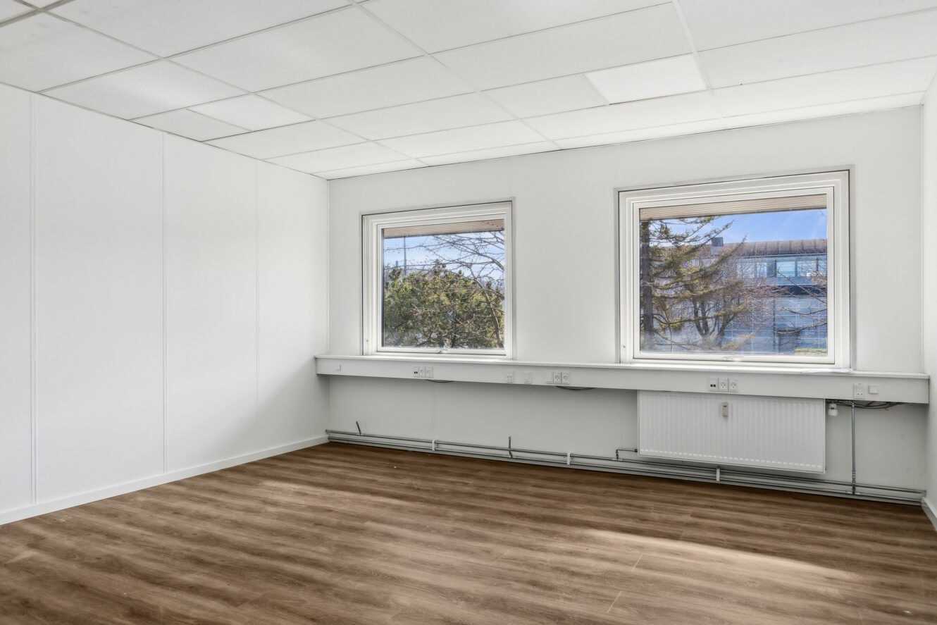Tomt kontorlokale med hvide vægge, to vinduer med udendørs udsigt, lyst trægulv og loftsplader. Langs væggen under vinduerne er der installeret stikkontakter og en radiator.