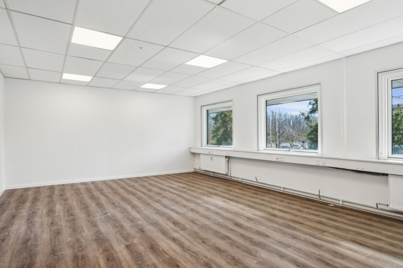 Tomt kontorlokale med hvide vægge, tre store vinduer, lysstofrør i loftet og vinylgulv i trælook.