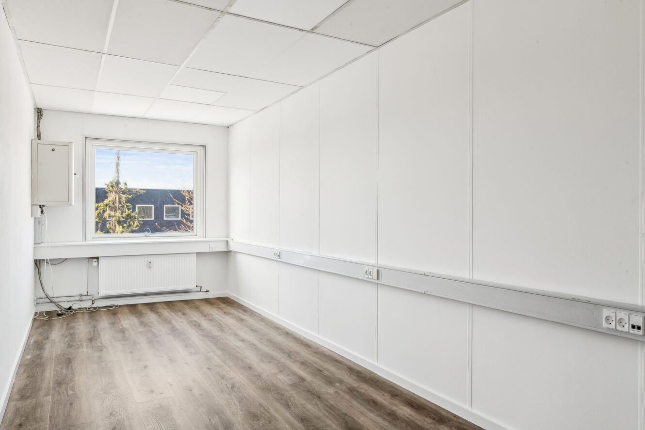 Et tomt, hvidt kontorlokale med træmønstret gulv, et vindue med udsigt til det fri, stikkontakter langs væggene og et loft med hvide fliser.