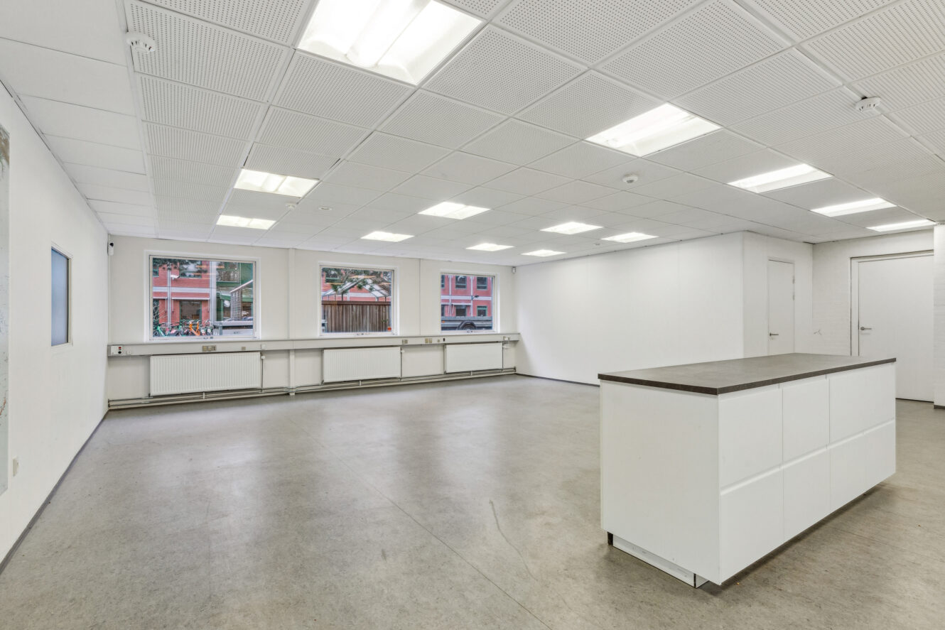 Rummeligt, tomt kontorlokale med hvide vægge, store vinduer, lysstofrør i loftet og en hvid midterskranke på et gråt gulv.