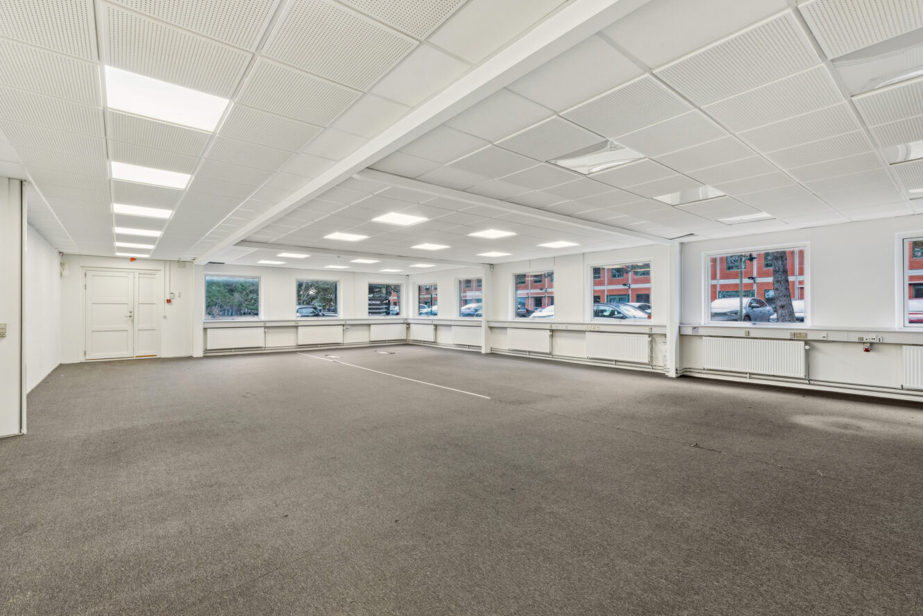 Rummeligt, tomt kontorlokale med gråt gulvtæppe, hvide vægge, store loftslamper og flere vinduer, der viser parkerede biler og nabobygninger udenfor.