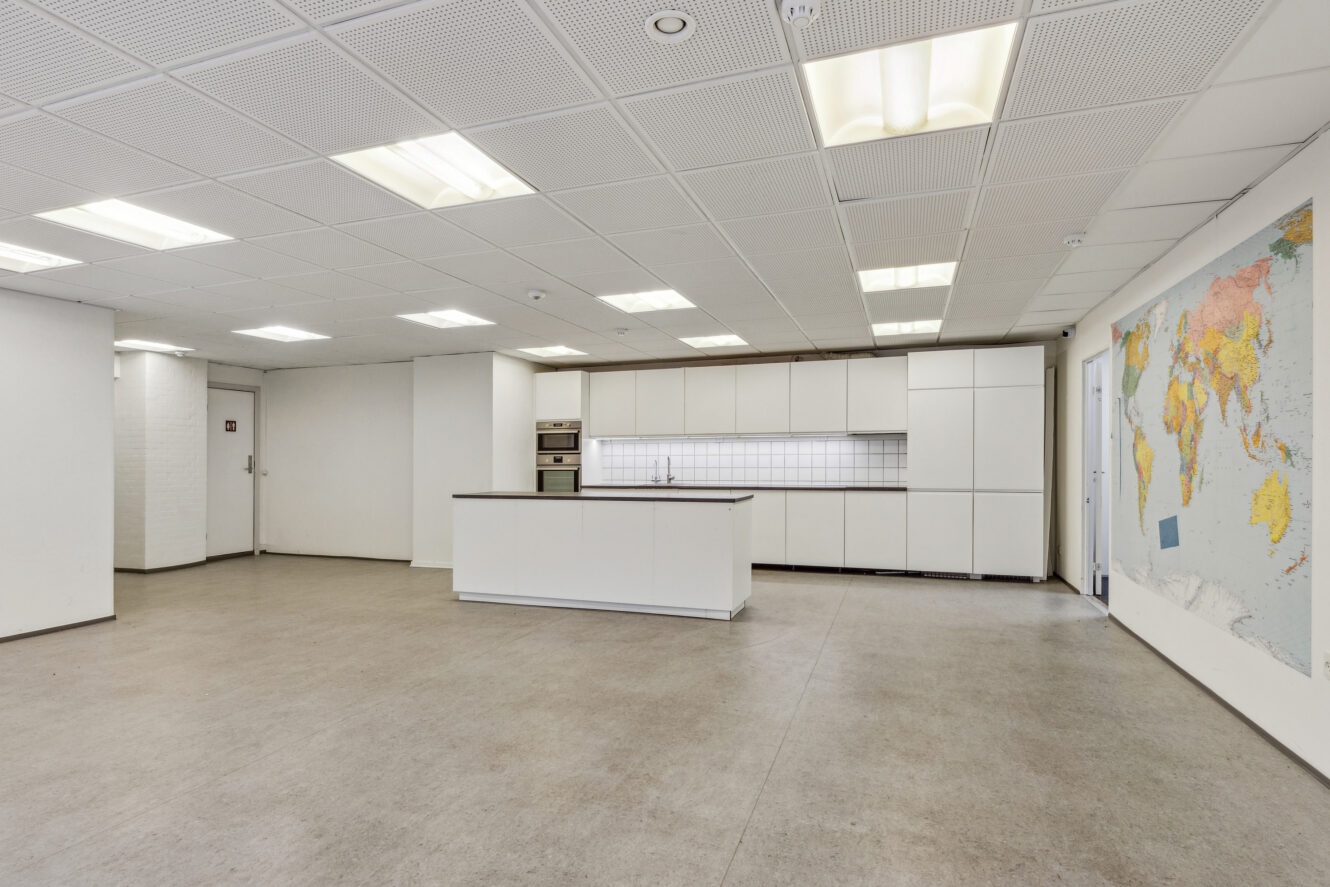 Rummeligt, moderne køkken med hvide skabe, ø, indbyggede ovne, flise-backsplash og et stort verdenskort på væggen i et lyst rum.