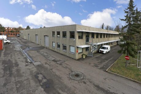 En toetagers industribygning med flere vinduer og et gråt ydre, en hvid varevogn parkeret ved indgangen og en stort set tom asfalteret plads omkring den.