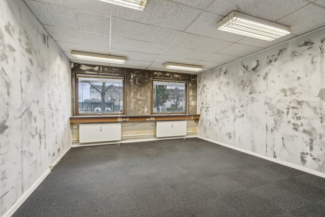 Tomt kontorlokale med plettede, ufærdige hvide vægge, gråt gulvtæppe, to vinduer, radiatorer nedenunder og lysstofrør i loftet.